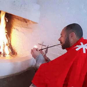 May include: A person wearing a bright red cape with a white cross emblem on the shoulder, holding a long, thin metal pipe near a lit fireplace. The individual has a beard and is positioned in profile, with a metal cross visible in the background.