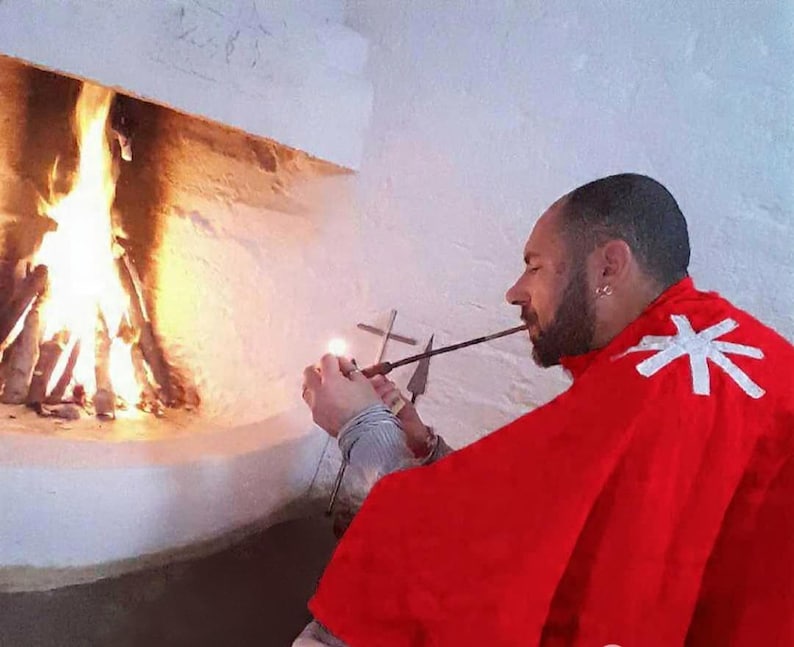 May include: A person wearing a bright red cape with a white cross emblem on the shoulder, holding a long, thin metal pipe near a lit fireplace. The individual has a beard and is positioned in profile, with a metal cross visible in the background.