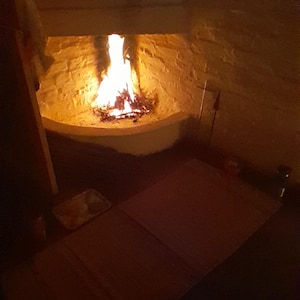 May include: A bright orange and yellow fire burns within a white stone corner fireplace. The flames illuminate the textured white brick walls of the hearth, casting a warm glow over the dark floor area in front of the fireplace opening.