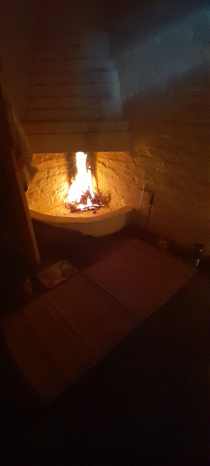 May include: A bright orange and yellow fire burns within a white stone corner fireplace. The flames illuminate the textured white brick walls of the hearth, casting a warm glow over the dark floor area in front of the fireplace opening.