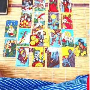 Op de afbeelding: Een verzameling van eenentwintig rechthoekige tarotkaarten gerangschikt in een rasterpatroon op een lichtgekleurd geweven oppervlak. Elke kaart bevat kleurrijke, gedetailleerde illustraties met diverse figuren, symbolen en scènes.