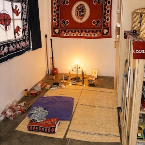 May include: A room featuring a wall tapestry with a central lion motif, a black metal candelabra with lit candles, two woven floor mats, a purple cushion, and various small objects arranged on the floor, including bowls and a framed photograph.