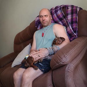 May include: A light blue sleeveless shirt, black patterned shorts, and a brown dapple dachshund. The person wears a red pendant necklace and multiple silver-toned bracelets on the left wrist, while resting on a brown leather recliner.