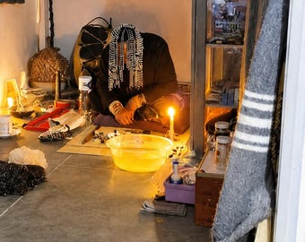 May include: A person with beaded hair sits on the floor in a dark room, wearing a black long-sleeved shirt. A yellow plastic basin filled with water sits in front of them, illuminated by a lit candle. A red tapestry with a wolf image hangs on the wall.