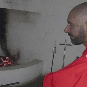 May include: A person with a shaved head and beard wearing a bright red garment sits before a stone fireplace with a small fire. A metal spear and a cross-shaped metal stand are positioned upright on the hearth near the flames.