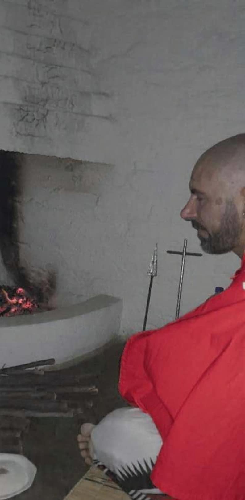 May include: A person with a shaved head and beard wearing a bright red garment sits before a stone fireplace with a small fire. A metal spear and a cross-shaped metal stand are positioned upright on the hearth near the flames.