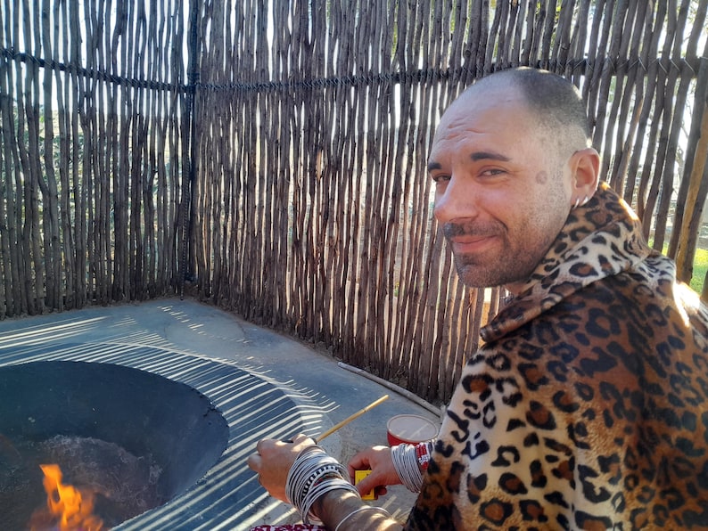 May include: A person wearing a leopard print garment and multiple silver-toned bracelets on their wrists, sitting near a circular fire pit. The individual has a short haircut and is holding a thin wooden stick near the flames.