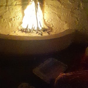 May include: A bright, flickering fire burns within a curved, light-colored stone fireplace. The flames cast a warm, golden glow against the textured masonry of the hearth, while the surrounding area remains in deep, dark shadow.