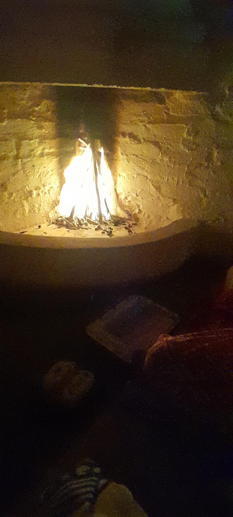 May include: A bright, flickering fire burns within a curved, light-colored stone fireplace. The flames cast a warm, golden glow against the textured masonry of the hearth, while the surrounding area remains in deep, dark shadow.