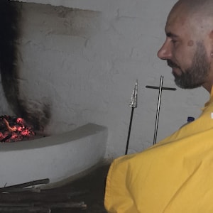 May include: A person with a shaved head and beard wearing a bright yellow robe and white garment with a black geometric pattern, sitting on a woven mat near a stone fireplace with glowing embers and a metal spear-like object nearby.
