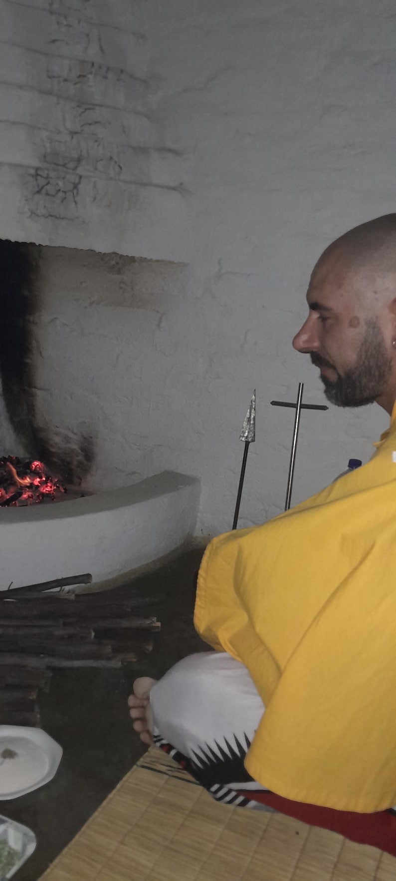 May include: A person with a shaved head and beard wearing a bright yellow robe and white garment with a black geometric pattern, sitting on a woven mat near a stone fireplace with glowing embers and a metal spear-like object nearby.