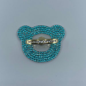 May include: A light blue phone ring holder with a gold band and a bear-shaped design. The ring holder is covered in rhinestones.