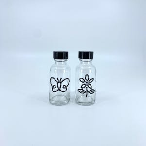May include: Two clear glass bottles with black lids. The bottle on the left has a black butterfly design and the bottle on the right has a black flower design.