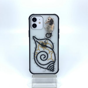 May include: A clear phone case with a black border, embellished with a seahorse and spiral design. The seahorse is gold and black, with a silver crystal cluster. The spiral and seahorse are adorned with black and gold crystals.