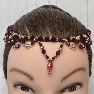 May include: A beaded headpiece with a burgundy and gold color scheme. The headpiece features multiple strands of burgundy beads and gold-colored accents. A central burgundy teardrop-shaped bead hangs from the center. The headpiece is designed to be worn on the forehead.