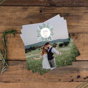 May include: Wedding invitation cards stacked on a wooden surface, featuring a couple in a field. The cards have a circular design with the words "We Do" and floral accents. A sprig of rosemary is placed on the wooden background.