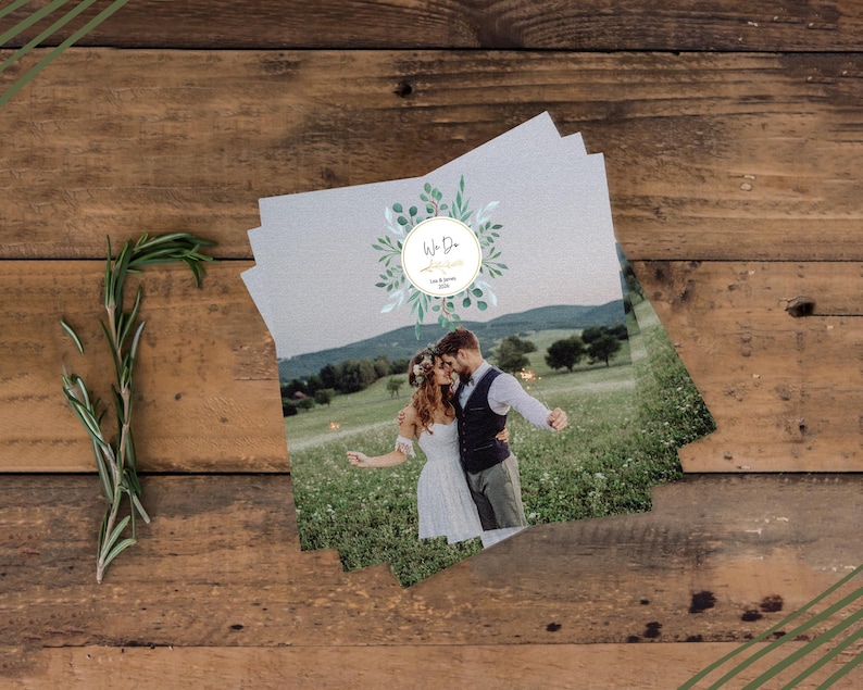 May include: Wedding invitation cards stacked on a wooden surface, featuring a couple in a field. The cards have a circular design with the words "We Do" and floral accents. A sprig of rosemary is placed on the wooden background.