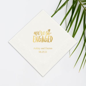 Op de afbeelding: Wit papieren servet met gouden letters die "We're so engaged" zeggen met een klein diamanten ringicoon. De tekst "Ashley and Darren 06.25.21" staat onder de hoofdtekst.