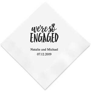 Op de afbeelding: Witte papieren servet met zwarte tekst die "we're so ENGAGED" luidt met een klein diamanten ringicoon. Onder de tekst staat "Natalie and Michael 07.12.2019".