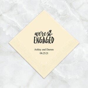 Op de afbeelding: Een cr&egrave;mekleurige papieren servet met zwarte tekst die "we're so ENGAGED" luidt met een klein diamanten ringicoon. Onder de tekst staat "Ashley and Darren 06.25.21".