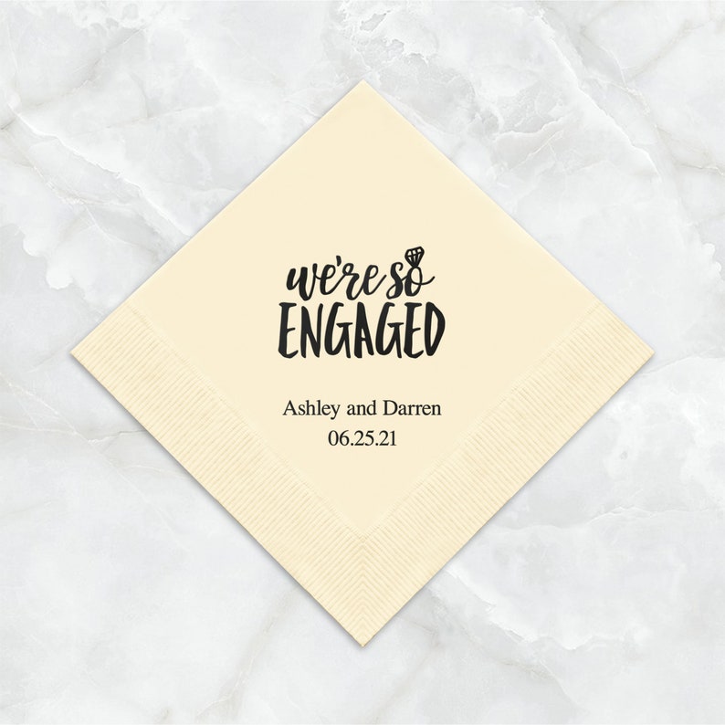 Op de afbeelding: Een cr&egrave;mekleurige papieren servet met zwarte tekst die "we're so ENGAGED" luidt met een klein diamanten ringicoon. Onder de tekst staat "Ashley and Darren 06.25.21".
