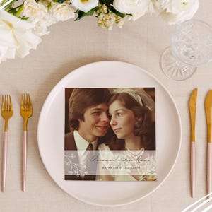May include: A square photo card with a vintage couple photo, text reading "Forever in Love" and "Happy 55th! 10.23.1970". The card sits on a white plate, with gold and pink cutlery, and a glass. White flowers are in the background.