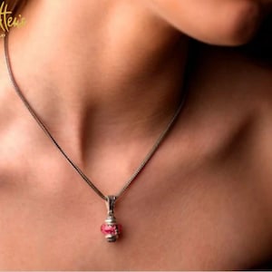 Puede incluir: Un collar de cadena de plata con un pequeño colgante que presenta una piedra preciosa rosa. El colgante tiene un diseño detallado y está engastado en un marco de plata.