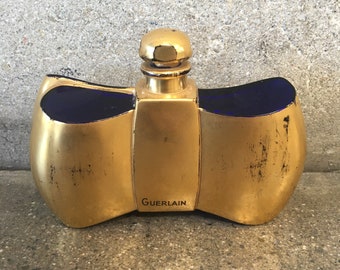 May include: A gold-colored perfume bottle with a bow-tie shape and a small, round stopper. The bottle has dark blue accents and the word "Guerlain" is printed on the front.