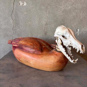 May include: A carved wooden sculpture of a bird-like form with a natural wood finish and red accents. A white animal skull is positioned at the front of the sculpture, with its jaws open. The sculpture is displayed on a dark surface.
