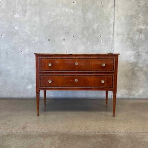 May include: A two-drawer wooden chest of drawers with a rich, warm brown finish. The chest features brass hardware and a contrasting dark wood inlay around the drawers. The piece has slender, tapered legs, and is set against a neutral background.
