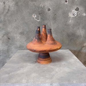 May include: A unique, handmade ceramic vase with a pedestal base. The vase features a wide, circular top with four cylindrical openings and a warm, earthy color palette of brown and black. The vase is displayed on a gray surface.