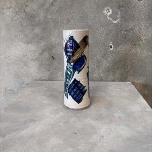 May include: A cylindrical ceramic vase with a cream-colored base. The vase is adorned with abstract brushstrokes in shades of blue, brown, and green. The design is applied in a seemingly random pattern, creating a unique and artistic look.