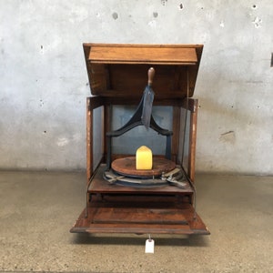 May include: Antique wooden cheese slicer with a black metal blade and a yellow block of cheese on the rotating platform. The top lid is open.