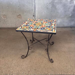 May include: Square side table with a colorful tile top featuring floral patterns in orange, blue, and yellow. The table has a black metal frame with decorative, scrolled legs. The table is approximately 20 inches tall.
