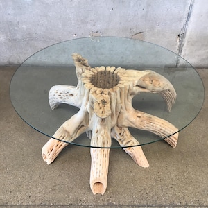 May include: A round glass top coffee table with a unique base made from a natural tree root. The base has a rough, textured surface and multiple branches extending out from the center.