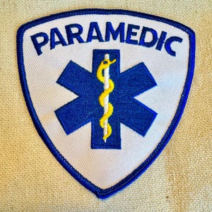 Könnte beinhalten: Weißer gestickter Aufnäher mit blauem Rand, der einen blauen Lebensstern mit einer gelben Schlange und einem Stab sowie das Wort "PARAMEDIC" in blauen Buchstaben zeigt.