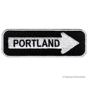 May include: Black and white embroidered patch with a white arrow pointing to the right and the word "PORTLAND" in white letters.