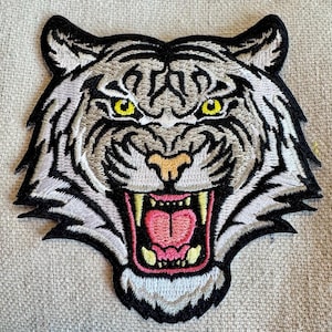 May include: White embroidered tiger head patch with black outline and yellow eyes. The tiger's mouth is open, showing sharp teeth and a pink tongue.