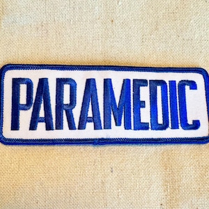 May include: Blue embroidered patch with a white background and the word "PARAMEDIC" in blue lettering.
