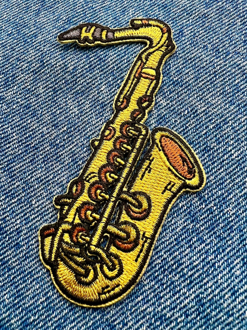 May include: A yellow embroidered saxophone patch with black and brown details. The patch is on a blue denim background.
