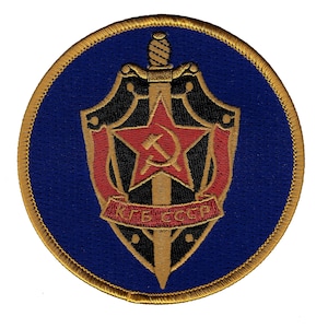 May include: A circular embroidered patch with a gold border and a blue background. The center of the patch features a black and gold shield with a red star, a hammer and sickle, and a sword. The text "KГБ СССР" is written on a red banner below the shield.