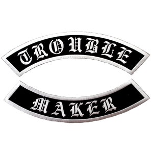 May include: Two black and white embroidered patches. The top patch says "TROUBLE" in white letters on a black background. The bottom patch says "MAKER" in white letters on a black background.