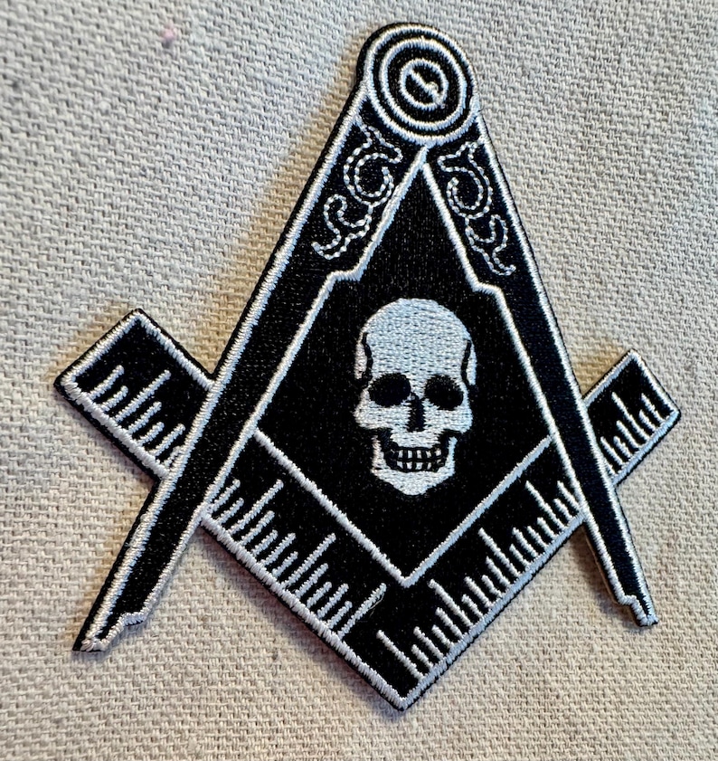 May include: Black and white embroidered patch of a skull inside a square and compass, a symbol of Freemasonry.