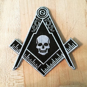May include: Black and white embroidered patch featuring a skull and crossbones inside a square and compass design. The patch is surrounded by a white border.