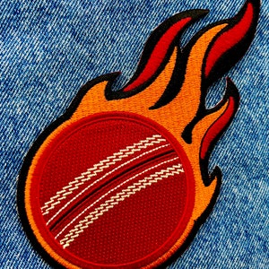 May include: A red and white cricket ball with black stitching is on fire. The flame is orange and yellow with black outlines. The image is on a blue denim background.