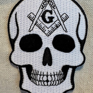 Könnte beinhalten: Weißer gestickter Totenkopf-Patch mit einem schwarzen freimaurerischen Symbol, das einen Zirkel und ein Geodreieck mit einem 'G' in der Mitte zeigt.