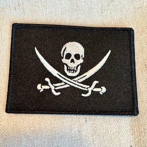 May include: A black fabric patch with a white embroidered skull and crossbones design. The skull has a detailed design with eye sockets, a nose, and a mouth.