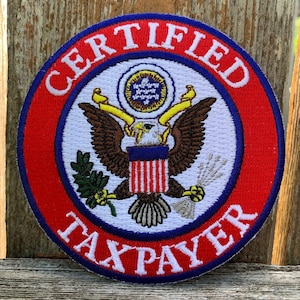 Peut inclure: Un écusson brodé rouge, blanc et bleu avec les mots "CERTIFIED TAXPAYER" autour du bord. Le centre de l'écusson représente un aigle avec un bouclier et une bannière.