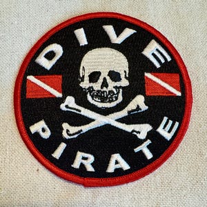 Può includere: Una toppa circolare nera e rossa con il testo "Dive Pirate" in lettere bianche attorno a un teschio e ossa incrociate bianche con una bandiera da sub rossa e bianca su entrambi i lati.