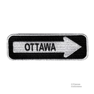 Puede incluir: Parche bordado en blanco y negro con una flecha blanca apuntando hacia la derecha y la palabra "OTTAWA" en letras negras.